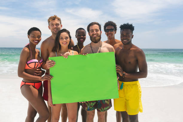 groupe multi-ethnique d'amis retenant une plaque verte vide à la plage par une journée ensoleillée - shirtless beach women bikini photos et images de collection