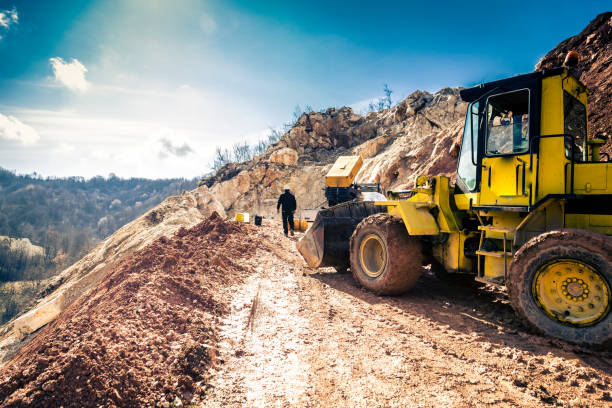 Mining worker working outdoors at the Quarry Wide angle color image depicting a mine working busy working outdoors at a limestone quarry. Rear view image of the worker while a yellow industrial digger is in the foreground. Bright blue sunny sky with cloudscape. Plenty of room for copy space. detonator stock pictures, royalty-free photos & images