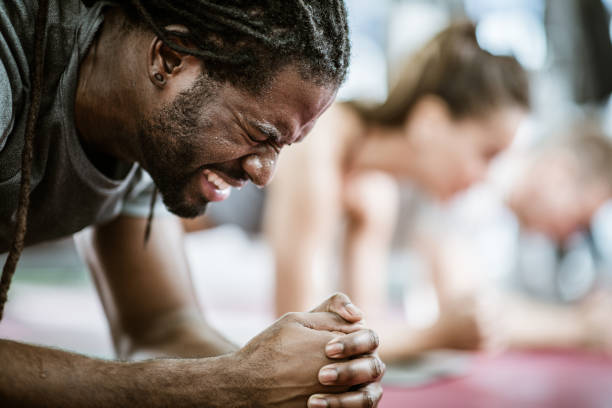 Exercising can be painful! Sweaty African American athlete making an effort while being in plank position in a health club. There are people in the background. effort stock pictures, royalty-free photos & images
