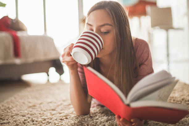 Girl relaxing at home with coffee and a book Young woman lying down on the floor in the bedroom, reading a book, relaxing. happy valentines day book stock pictures, royalty-free photos & images