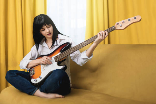 bonita mujer joven teniendo sus piernas cruzadas tocando algún disco o ejercicio en el bajo de la guitarra sentado en el sofá en el estilo de vida casero - bass guitar fotografías e imágenes de stock