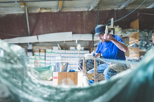 An Alaskan fisherman prepares and checks gill nets in his work space in a crowded warehouse area. He is sitting down and has the net spread out in front of him.