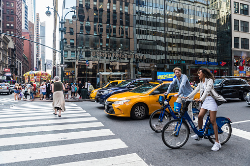 New York City, USA - July 28, 2018: Cyclists couple with Citibike bicycles on Fifth Avenue (5th Avenue) with people around and traffic in Midtown Manhattan, New York City, USA
