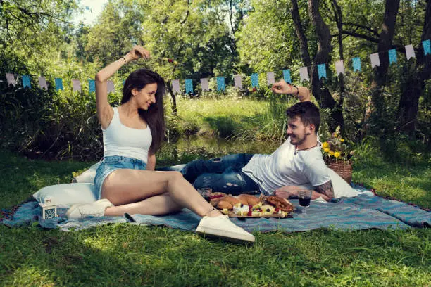 Photo of Young couple on a picnic in a city park sitting on a blanket and dueling with skewers with fruits Selective focus