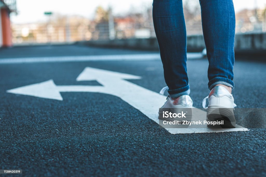 Make decision which way to go. Walking on directional sign on asphalt road. Female legs wearing jeans and white sneakers. Direction Stock Photo