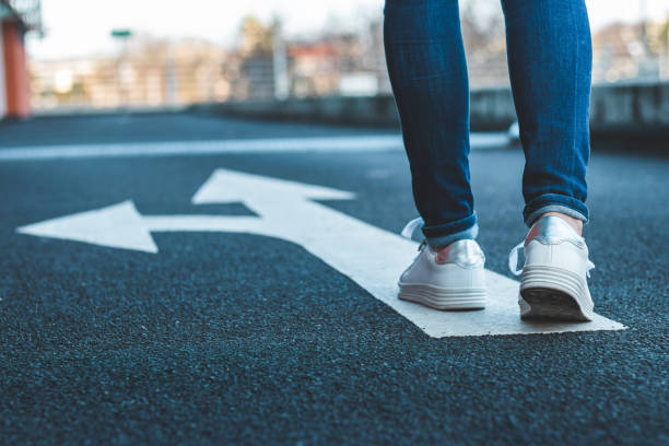Make decision which way to go. Walking on directional sign on asphalt road. Female legs wearing jeans and white sneakers. guidance stock pictures, royalty-free photos & images
