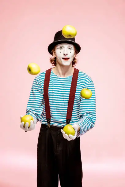Pantomime with white facial makeup juggling with apples on the pink background in the studio