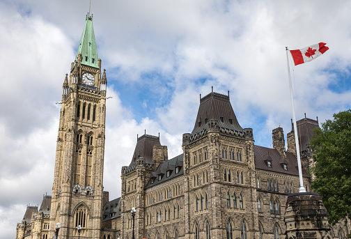 A Panoramic Shot Of Canadian Parliament Building In Ottawa Canada With Blue Skies And Clouds Behind And Canadian Flag Blowing In The Wind