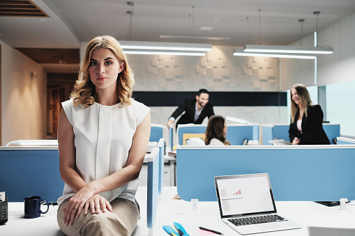 Portrait of serious female employee excluded by colleagues. Blonde businesswoman sitting on desk at work in modern coworking space, looking at camera with sad expression.