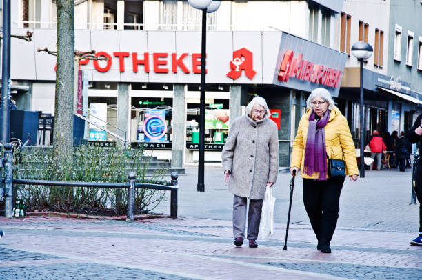 fora para um passeio à noite. duas mulheres idosas (incl. uma com a ajuda de um bastão) que anda em um dia de inverno fora de uma farmácia alemão local (lowen apotheke) em hombruch centrum, dortmund-alemanha - german culture people women germany - fotografias e filmes do acervo