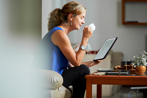 Mature woman resting at home, she sitting on sofa in front of the table drinking coffee and watching something on tablet pc