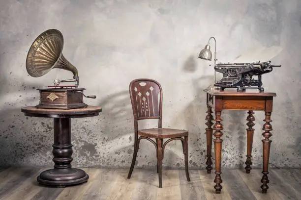 Photo of Vintage loft room with old typewriter and lamp on wooden desk, antique chair and classic gramophone with brass horn front concrete wall background with shadows. Retro style filtered photo