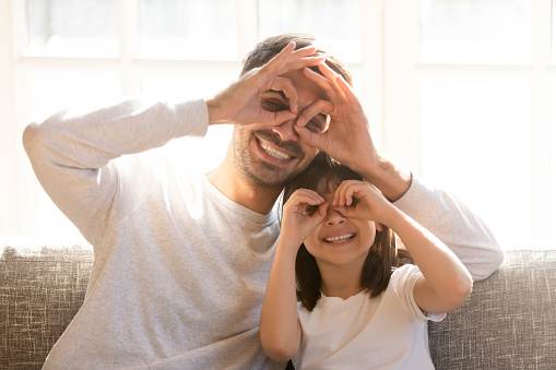Pretty family father adorable daughter sitting on sofa do funny faces making with fingers eyewear shape like glasses looking through binoculars, have fun with child free time play tricks fool concept