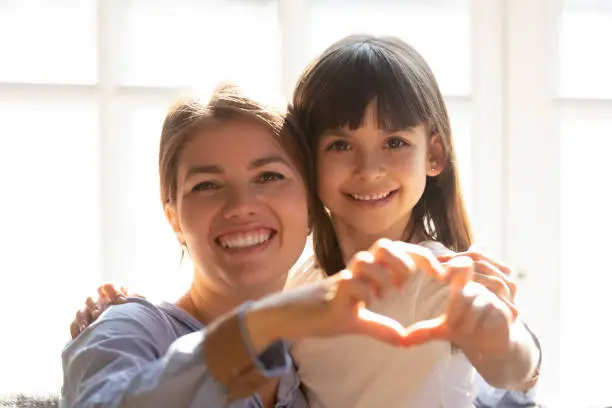 Head shot portrait mother and daughter make heart shape with hands fingers hugging sitting on couch sunlight through the window. Adoption and custody, health medical pediatric insurance, love concept