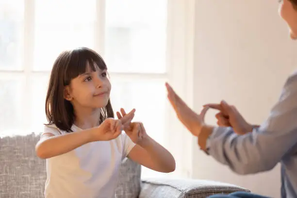 Mom communicating with deaf daughter focus on kid sitting on couch in living room make fingers shape hands talking nonverbal. Hearing loss deaf disability person sign language learning school concept