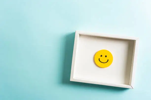 Photo of Concept of well-being, well done, feedback, employee recognition award. Happy yellow smiling emoticon face frame hanging on blue background with empty space for text.