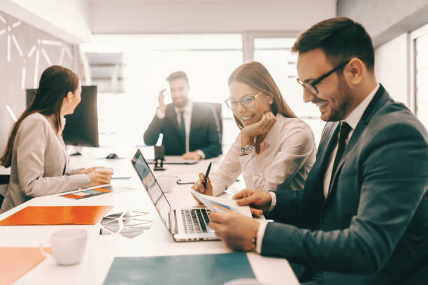 istock Two cheerful colleagues in formal wear solving problem and looking at paperwork. In background employees talking. It is literally true that succeed best and quickest by helping others to succeed. 1138482589