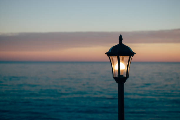 Modern coastal lamp post standing among seaside rocks with waves crashing in the background. Urban and nature contrast captured at a clear, sunny waterfront.