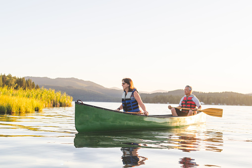 A Caucasian couple spend a summer evening canoeing around a beautiful lake as the sun sets in the background. There are mountains and forested areas in the background.