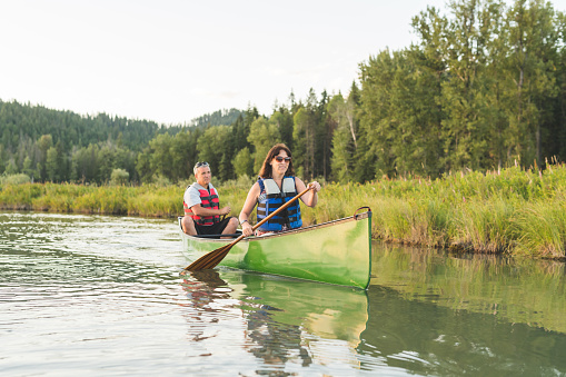 A Caucasian couple spend a summer afternoon canoeing around a beautiful lake. There are mountains and forest in the background and the sun is setting on a lovely day. She is in front as they paddle from left to right.