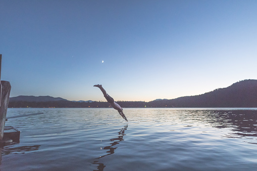 A Caucasian male dives in the lake from the diving board on the pier. It is dusk. On the other side of the lake are houses, forest, and a mountain range. He is suspended over the water and the moon is visible in the sky.