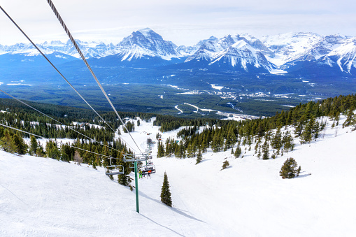 Unidentifiable skiers on chairlift going up a ski slope in the snowy mountain range of the Canadian Rockies.