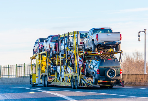 Big rig industrial grade yellow classic bonnet car hauler semi truck transporting cars on two levels semi trailer driving on the overpass road with protective fence on the side