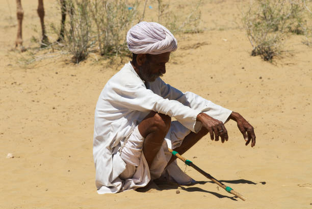 Jamba, India - April 02, 2007: Unidentified camel herder wearing wight clothes sits on the sand in Thar desert in Jamba, India.