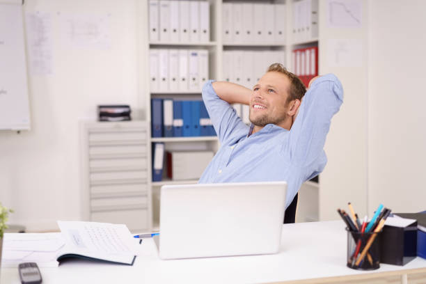 A young man leans back in his chair with hands behind his head, gazing upward in thought. He is daydreaming in the office.