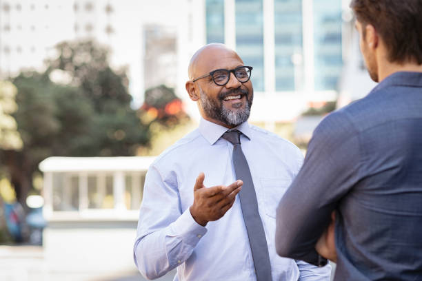 Business people talking outdoor Smiling mature african businessman talking to his colleague outside the office. Two cheerful business men talking to each other and gesturing. Mature bald man with beard and spectacles in conversation with young colleague on street. businessman happiness outdoors cheerful stock pictures, royalty-free photos & images