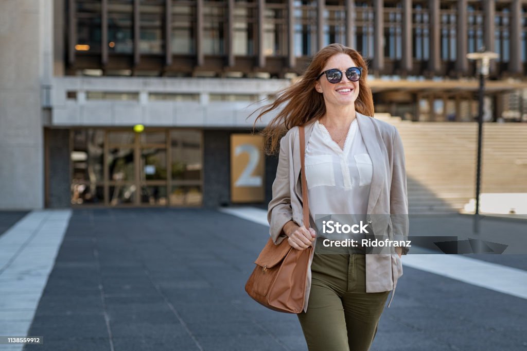 Happy mature woman walking confidently Portrait of successful happy woman on her way to work on street. Confident business woman wearing blazer carrying side bag walking with a smile. Smiling woman wearing sunglasses and walking on city street. Women Stock Photo