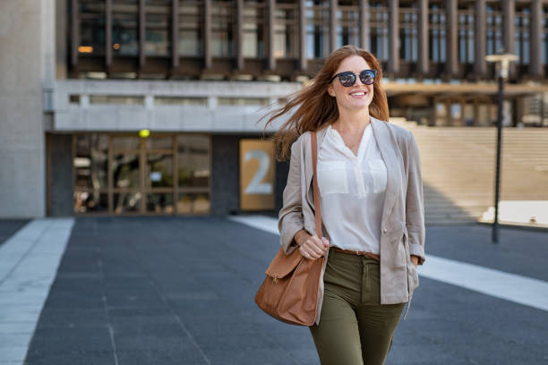 Happy mature woman walking confidently Portrait of successful happy woman on her way to work on street. Confident business woman wearing blazer carrying side bag walking with a smile. Smiling woman wearing sunglasses and walking on city street. business casual fashion stock pictures, royalty-free photos & images