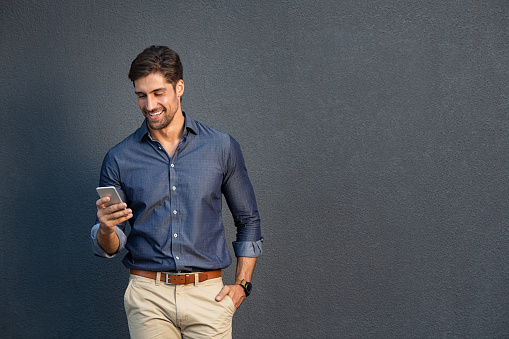 Portrait of young man leaning against a grey wall using mobile phone. Happy business man messaging with smartphone isolated on gray background with copy space. Smiling casual guy typing and reading a message on cellphone leaning on wall.