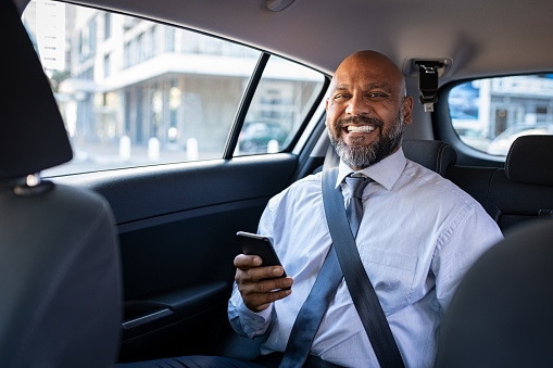 African mature businessman holding smartphone while looking at camera. Successful entrepreneur wearing blue tie sitting in car using mobile phone. Happy formal black business man traveling in taxi.