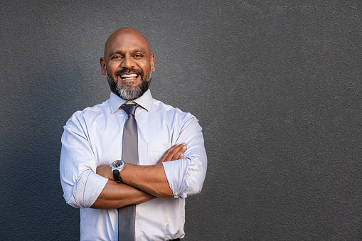 Successful senior man with folded arms standing over grey background. Handsome mature black businessman in shirt and tie looking at camera. Portrait of joyful business man on a grey wall with copy space.