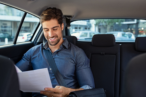 Business man looking at documents with concentration in his car. Young happy businessman reading contract on car back seat and smiling. Successful entrepreneur reading paperwork documents while sitting in taxi.