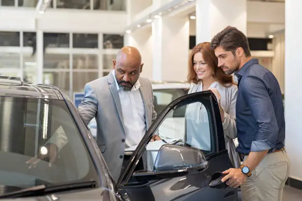 Mature african salesman showing all the car features to young couple. Car dealer showing new car to young man and beautiful woman in showroom. Couple buying a new suv in a auto dealership.
