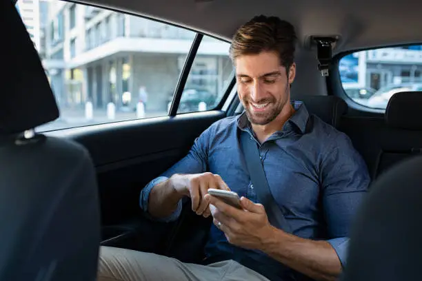 Happy smiling businessman man typing message on phone while sitting in a taxi. Young businessman in formal clothing using smartphone while sitting on back seat in car. Cheerful guy messaging wirth cellphone.