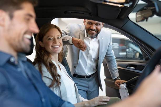 Salesman showing new car to couple African salesman showing new car to young couple in showroom. Happy man and woman choosing auto in car dealership while cardealer explaining the new technology. Smiling beautiful couple test the comfort of their new suv. car salesperson photos stock pictures, royalty-free photos & images