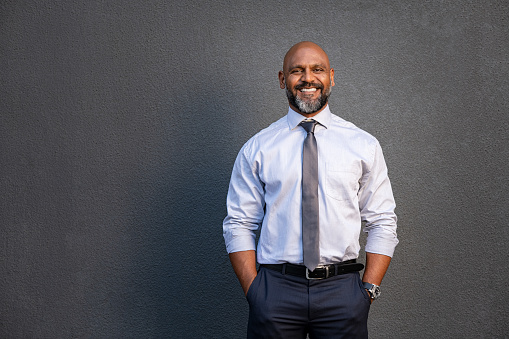 Portrait of successful black businessman standing against grey wall. Handsome senior entrepreneur wearing blue tie and formal clothing smiling and looking at camera. Mature happy man with hands in pocket isolated on grey background with copy space.