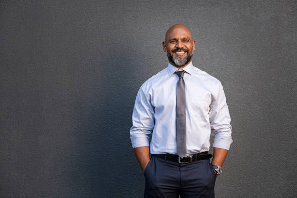 African american businessman smiling on grey Portrait of successful black businessman standing against grey wall. Handsome senior entrepreneur wearing blue tie and formal clothing smiling and looking at camera. Mature happy man with hands in pocket isolated on grey background with copy space. professional portrait stock pictures, royalty-free photos & images