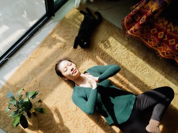 Photo of Pretty Chinese young woman meditating at home, lying on floor with her black cat in sun light, exercise, Lotus pose, prayer position, namaste, working out, Feeling peace and wellness concept.