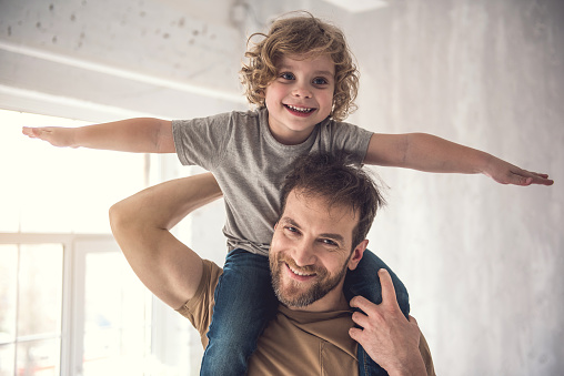 Happy little boy is piggybacking with his dad indoors. Cheerful parent is holding child while kid is putting up arms like plane and looking playfully at camera