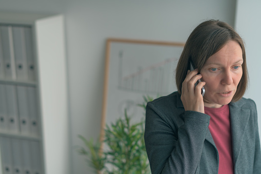 Businesswoman in mobile phone conversation by the office window, adult female caucasian business person talking on smartphone