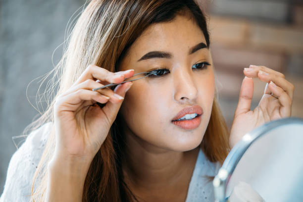 Asian woman placing decorative eyelashes on eye Closeup young Asian model placing decorative eyelashes on eye on blurred background false lashes stock pictures, royalty-free photos & images