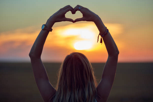 menina que prende o símbolo da coração-forma para o amor no por do sol/nascer do sol tempo. - heart shape loneliness women praying - fotografias e filmes do acervo