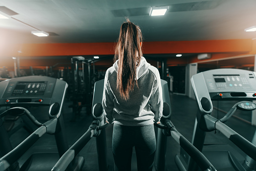 Slim brunette in sweatshirt and ponytail standing on treadmill in gym at night. Backs turned. Do your best and forget the rest.