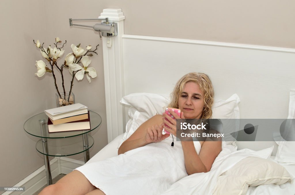 Woman using cell phone Blonde woman relaxing on a bed after a bath and using cell phone Adult Stock Photo