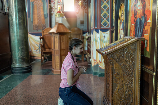 Beautiful Young Female Teenager is Standing by the Altar Inside the Church, Surrounded by Holy Icons and is Praying to the God. Young Schoolgirl with Long Hair and Backpack is Talking to the God While Kneeling in Church Interior near the Holy Icon. Faith, Spirituality and Religion Concept.