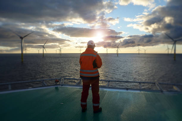 Offshore manual worker standing on helipad with wind-turbines behind him in sunset Wind-turbine, offshore, worker, climbing, sun, big, deck, vessel offshore platform stock pictures, royalty-free photos & images